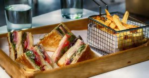 a toasted sandwich on a wooden tray with a metal basket of fries on the side